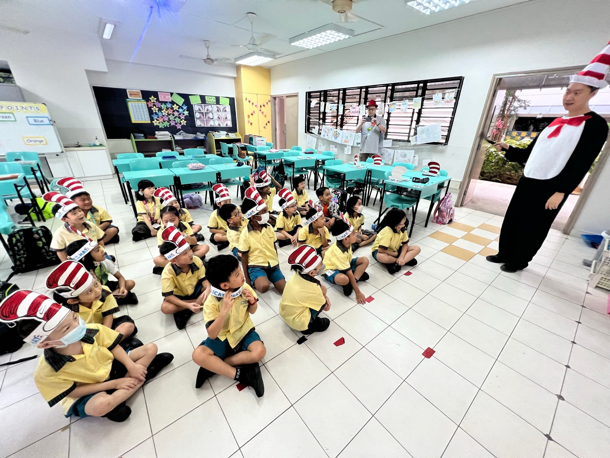 Children in yellow uniforms and red-striped Cat in the Hat paper hats sit on a classroom floor facing a costumed adult.
